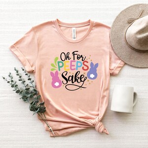 May include: A pink t-shirt with the text "Oh For Peeps Sake" in colorful letters and a cartoon bunny graphic.