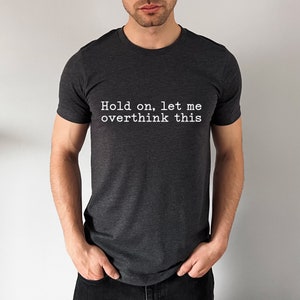 May include: A dark grey t-shirt with the text "Hold on, let me overthink this" printed in white.