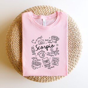 May include: A light pink t-shirt with a black graphic design featuring the word "Scorpio" and related illustrations. The design includes a constellation, a teacup, and other symbols. The shirt is displayed on a woven, circular surface.