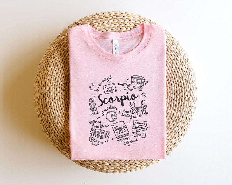 May include: A light pink t-shirt with a black graphic design featuring the word "Scorpio" and related illustrations. The design includes a constellation, a teacup, and other symbols. The shirt is displayed on a woven, circular surface.