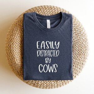 Pu&ograve; includere: Una maglietta grigia scura con la scritta "Easily Distracted by Cows" stampata in bianco.