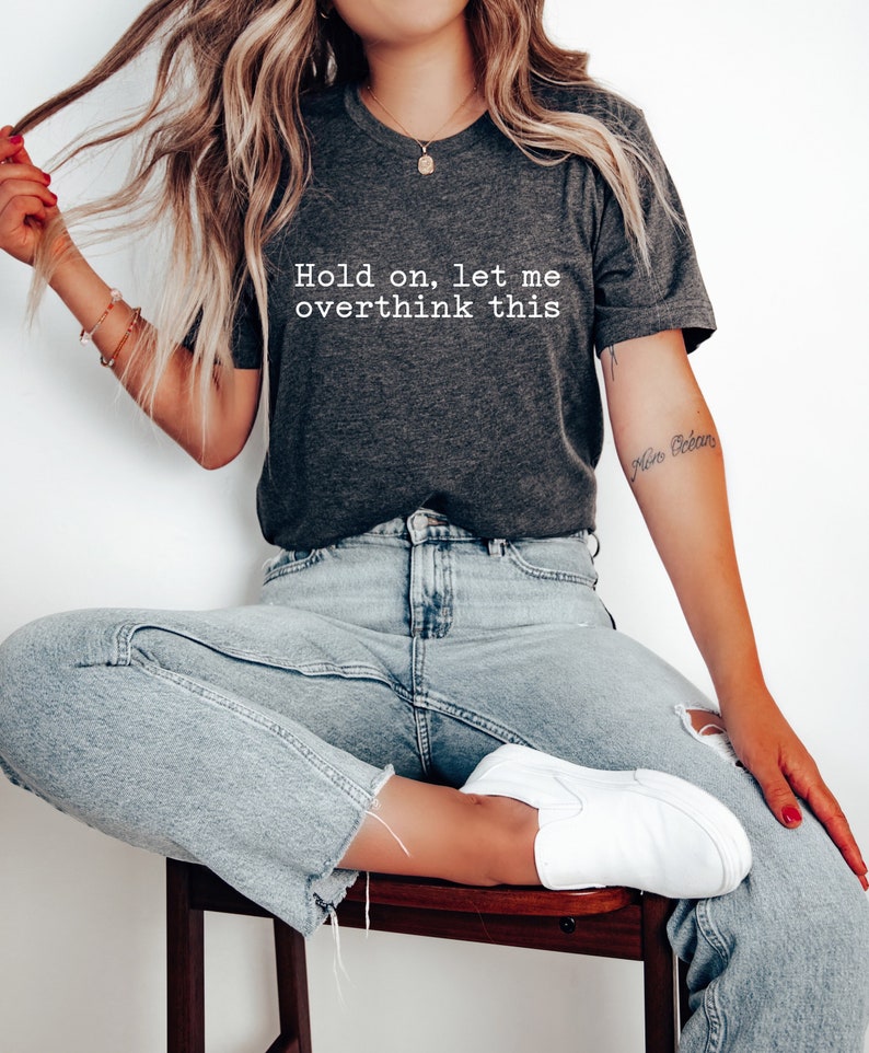 May include: A dark gray t-shirt with the text "Hold on, let me overthink this" printed in white. The model is wearing the shirt with light blue jeans and white sneakers.