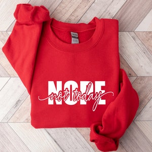 Puede incluir: Una sudadera roja brillante con la palabra "NOPE" en letras blancas grandes y las palabras "not today" en una fuente cursiva. La sudadera est&aacute; doblada, con las mangas visibles.