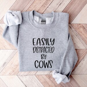 Pu&ograve; includere: Felpa grigia con la scritta "EASILY DISTRACTED BY COWS" stampata in nero.