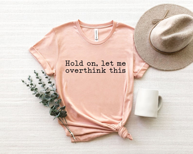 May include: A peach-colored t-shirt with the text "Hold on, let me overthink this" printed in black.