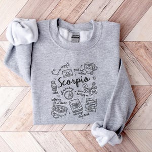 May include: A light gray crewneck sweatshirt featuring a black graphic design related to the Scorpio zodiac sign. The design includes the word "Scorpio" and various illustrations such as a constellation, a cup of coffee, and a book.