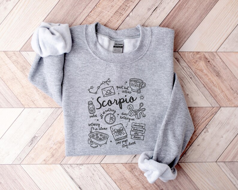 May include: A light gray crewneck sweatshirt featuring a black graphic design related to the Scorpio zodiac sign. The design includes the word "Scorpio" and various illustrations such as a constellation, a cup of coffee, and a book.