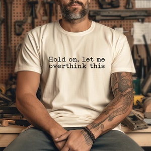 May include: A white t-shirt with the text "Hold on, let me overthink this" printed on the front. The person wearing the shirt has a beard and tattoos on their arm.