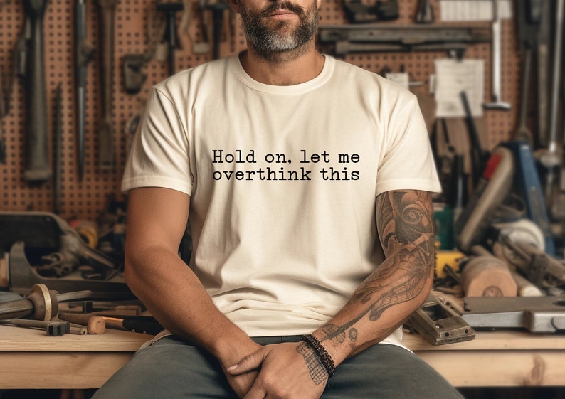 May include: A white t-shirt with the text "Hold on, let me overthink this" printed on the front. The person wearing the shirt has a beard and tattoos on their arm.