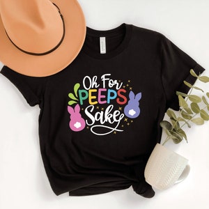 May include: Black t-shirt with a colorful graphic that says "Oh For Peeps Sake" with two pink and purple bunny-shaped marshmallow candies.