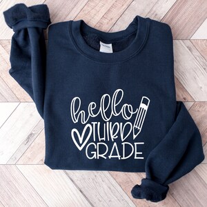 Puede incluir: Una sudadera azul marino con el texto "hello third grade" en blanco, con un coraz&oacute;n y un l&aacute;piz.
