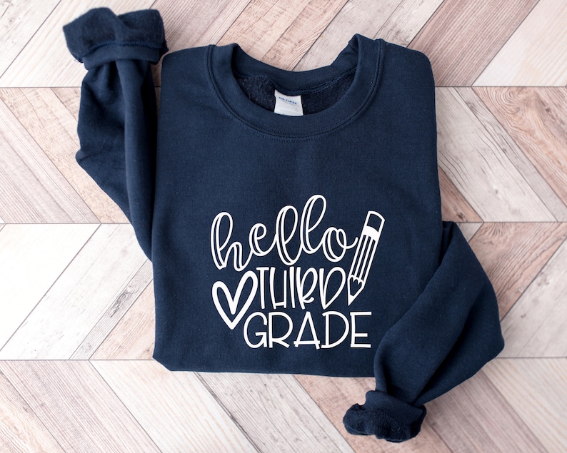 Puede incluir: Una sudadera azul marino con el texto "hello third grade" en blanco, con un coraz&oacute;n y un l&aacute;piz.