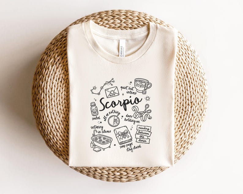 May include: Cream-colored t-shirt with a black graphic design featuring the word "Scorpio" and related illustrations. The design includes a constellation, envelope, teacup, and other symbols. The shirt is folded on a woven surface.