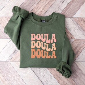 May include: An olive green sweatshirt with the word "DOULA" in a retro font, repeated three times in shades of pink and orange. The sweatshirt is folded neatly on a light wood-grain surface.