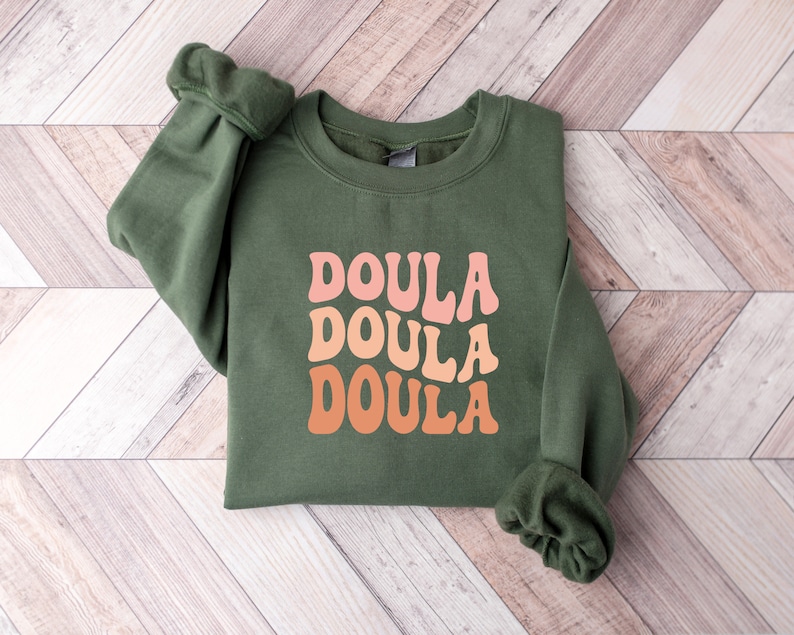 May include: An olive green sweatshirt with the word "DOULA" in a retro font, repeated three times in shades of pink and orange. The sweatshirt is folded neatly on a light wood-grain surface.