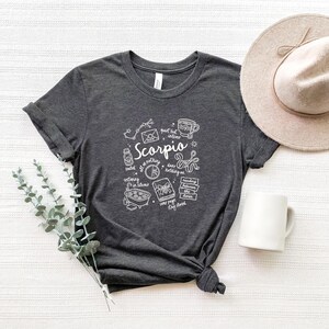 May include: Dark gray t-shirt with a white graphic design featuring the word "Scorpio" and related symbols. A beige hat and white mug are next to the shirt. A sprig of green leaves is on the left.