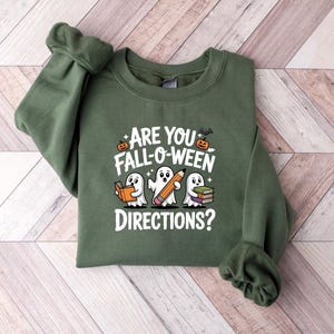 May include: An olive green sweatshirt with the text "ARE YOU FALL-O-WEEN DIRECTIONS?" and illustrations of ghosts, a pencil, books, a pumpkin, and a bat. The sweatshirt is folded on a light wood-grain surface.