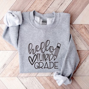 Puede incluir: Sudadera gris con el texto "hello third grade" en letras negras con un coraz&oacute;n y un gr&aacute;fico de l&aacute;piz.