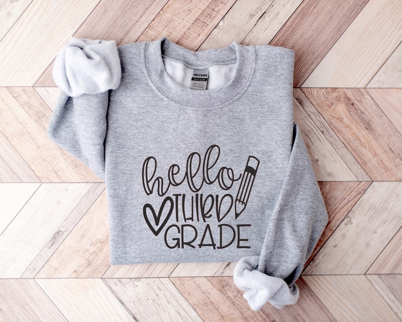 Puede incluir: Sudadera gris con el texto "hello third grade" en letras negras con un coraz&oacute;n y un gr&aacute;fico de l&aacute;piz.