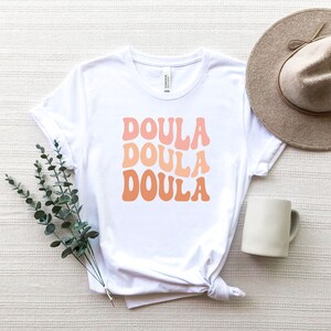 May include: White t-shirt with the word "DOULA" in three stacked lines, each in a different shade of orange. A beige hat and a white mug are next to the shirt. A sprig of green leaves is placed near the shirt.