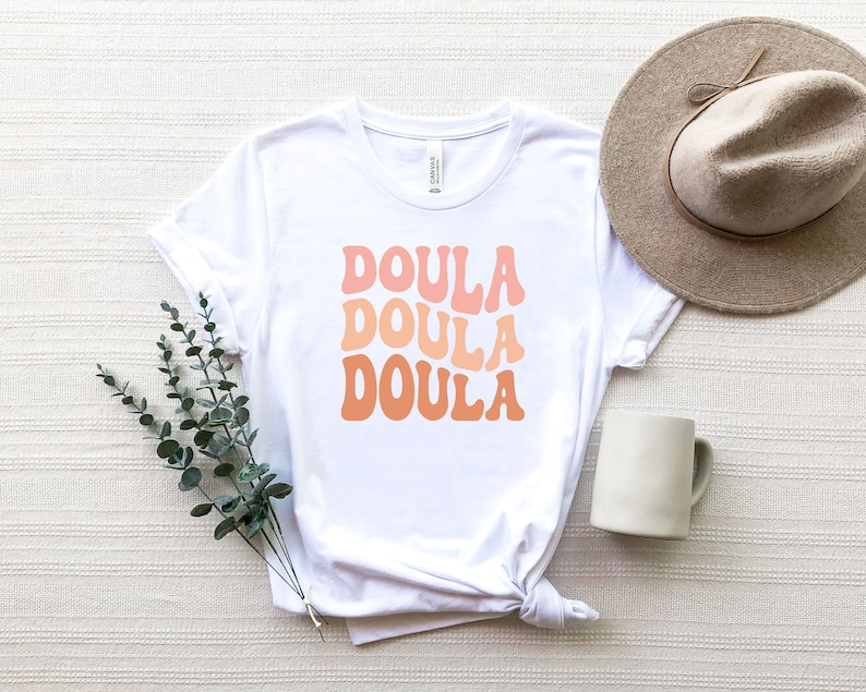 May include: White t-shirt with the word "DOULA" in three stacked lines, each in a different shade of orange. A beige hat and a white mug are next to the shirt. A sprig of green leaves is placed near the shirt.