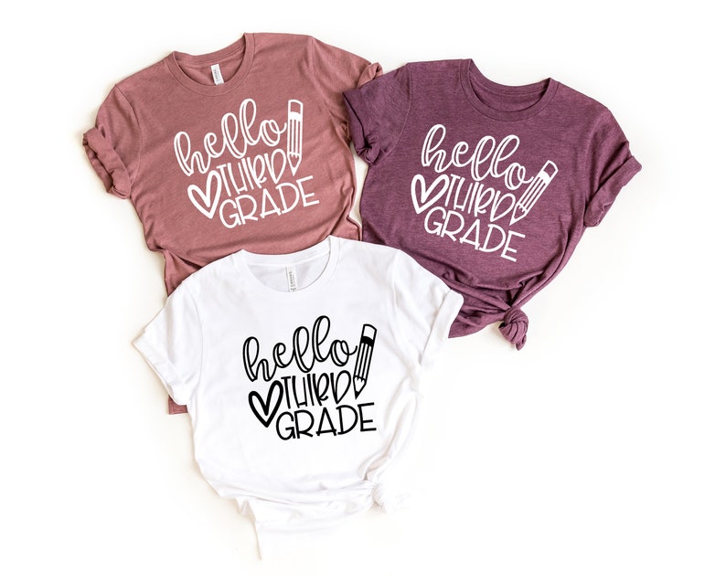 Puede incluir: Tres camisetas con el texto "hello third grade" en letras blancas con un l&aacute;piz negro y un dise&ntilde;o de coraz&oacute;n. Una camiseta es blanca, otra es malva y otra es de un tono malva m&aacute;s oscuro.