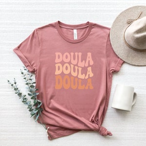 May include: A dusty rose t-shirt with the word "DOULA" in a retro font stacked three times in shades of peach and orange. A beige hat and white mug are next to the shirt. A sprig of green leaves is on the left.