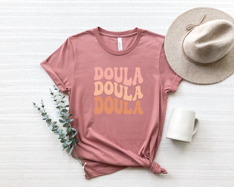May include: A dusty rose t-shirt with the word "DOULA" in a retro font stacked three times in shades of peach and orange. A beige hat and white mug are next to the shirt. A sprig of green leaves is on the left.