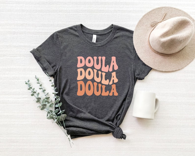May include: Dark gray t-shirt with the word "DOULA" in a retro font, stacked three times in shades of pink and orange. A beige hat and white mug are next to the shirt. Eucalyptus sprigs are in the lower left corner.