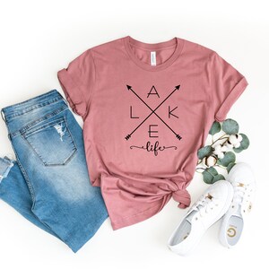 Op de afbeelding: Een stoffig roze t-shirt met de woorden "LAKE life" in zwart, gekruiste pijlen en een cursief lettertype. Het shirt wordt gecombineerd met lichtblauwe jeans, witte sneakers en takjes groen. Het shirt is in de taille geknoopt.