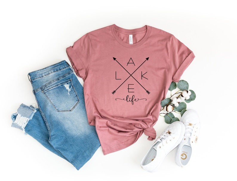 Op de afbeelding: Een stoffig roze t-shirt met de woorden "LAKE life" in zwart, gekruiste pijlen en een cursief lettertype. Het shirt wordt gecombineerd met lichtblauwe jeans, witte sneakers en takjes groen. Het shirt is in de taille geknoopt.