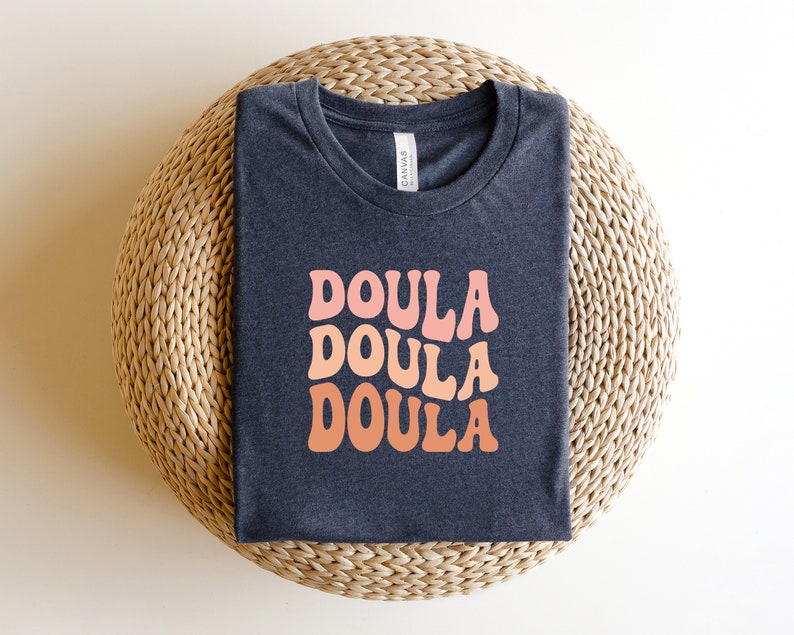 May include: A navy blue t-shirt with the word "DOULA" in a retro font, repeated three times in shades of pink and orange. The shirt is folded neatly on a woven, circular surface.