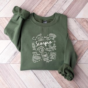 May include: A green crewneck sweatshirt with white graphic illustrations and text. The design includes the word "Scorpio" with related symbols and phrases. The sweatshirt is folded on a wooden surface.