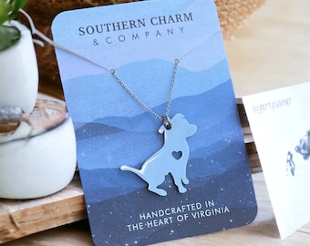 May include: A silver-toned metal necklace featuring a pendant shaped like a sitting dog with a small heart-shaped cutout. The necklace is displayed on a blue card printed with the text Southern Charm & Company and Handcrafted in the Heart of Virginia.