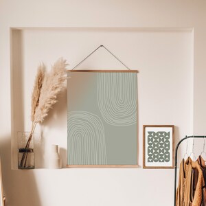 May include: Abstract art print in sage green and white, framed in wood, hanging on a wall. The design features two curved white lines. A smaller framed print with a geometric pattern is also visible.