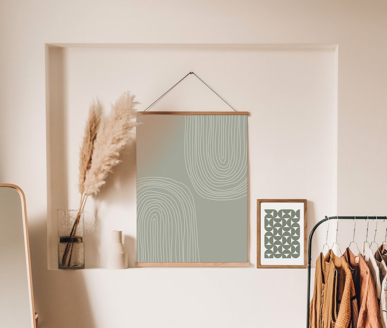 May include: Abstract art print in sage green and white, framed in wood, hanging on a wall. The design features two curved white lines. A smaller framed print with a geometric pattern is also visible.