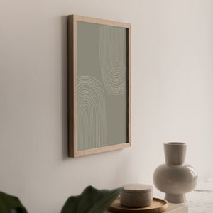 May include: A framed print with a minimalist design of two arched shapes in white lines on a muted green background. The artwork is displayed on a wall, with a beige vase, a small round container, and a wooden tray.