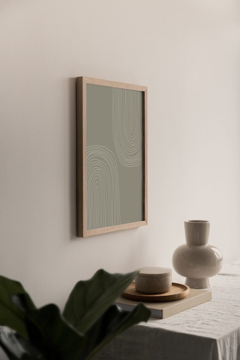 May include: A framed print with a minimalist design of two arched shapes in white lines on a muted green background. The artwork is displayed on a wall, with a beige vase, a small round container, and a wooden tray.