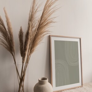 May include: A framed print with a sage green background and white arched lines. A clear glass vase holds dried pampas grass, and a small, beige ceramic vase sits nearby. The scene evokes a minimalist, bohemian aesthetic.