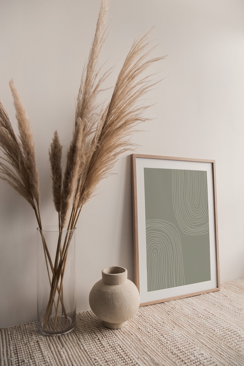 May include: A framed print with a sage green background and white arched lines. A clear glass vase holds dried pampas grass, and a small, beige ceramic vase sits nearby. The scene evokes a minimalist, bohemian aesthetic.