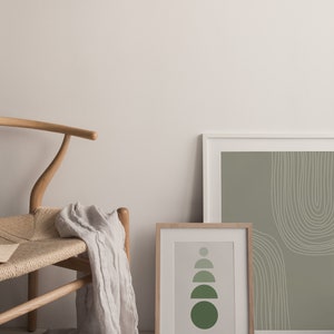May include: Two framed art prints with abstract designs. One print features a series of green semi-circles, the other has white curved lines on a green background. A wooden chair with a woven seat and a draped linen scarf complete the scene.