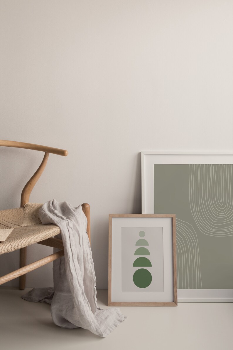 May include: Two framed art prints with abstract designs. One print features a series of green semi-circles, the other has white curved lines on a green background. A wooden chair with a woven seat and a draped linen scarf complete the scene.