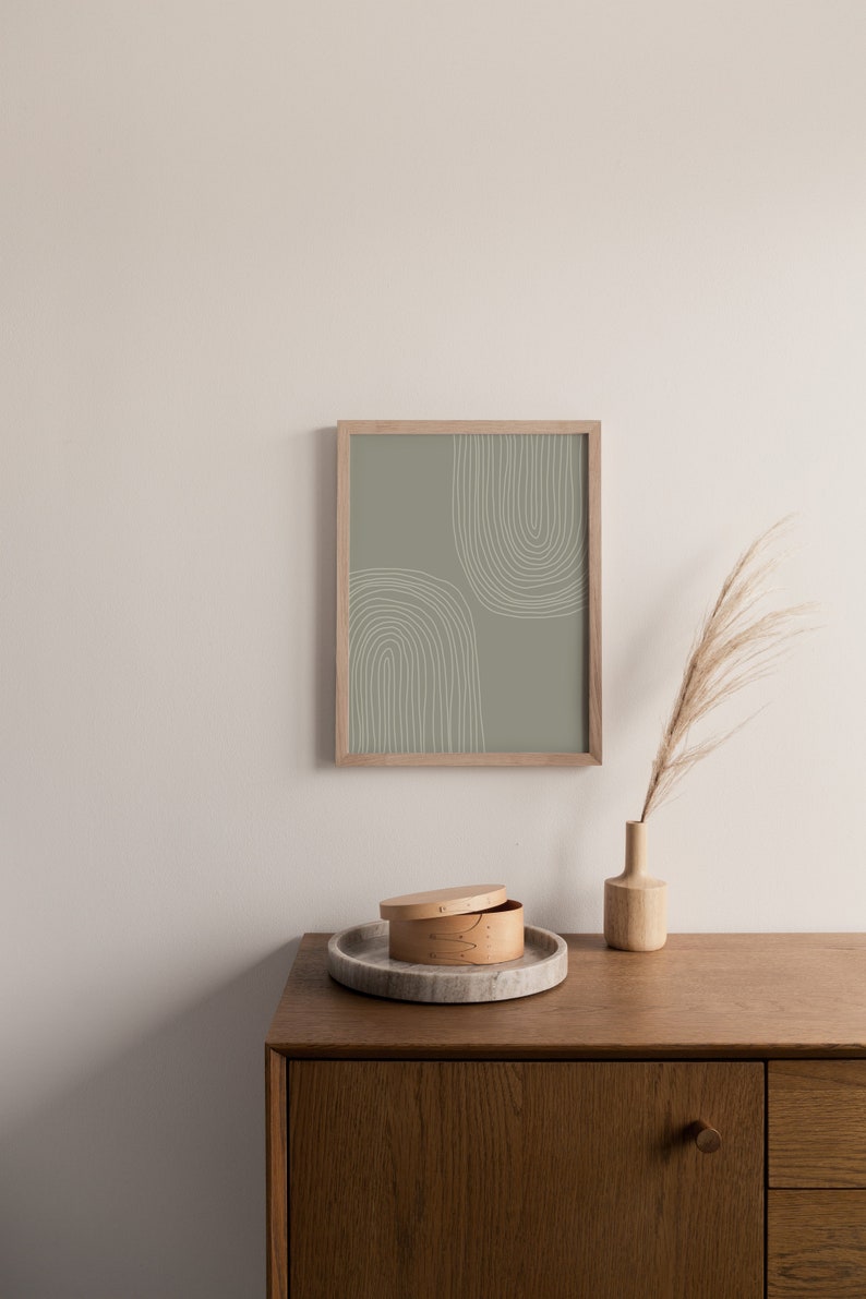 May include: A framed print with a sage green background and white arched lines. A wooden vase with dried grass and a round wooden box with a lid sit on a wooden cabinet. A marble tray holds the box.