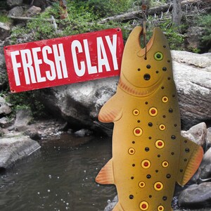 May include: A brown and yellow ceramic fish with black and red spots hanging from a hook. The fish is hanging over a stream with rocks and green foliage. A red sign with white letters reads "FRESH CLAY".