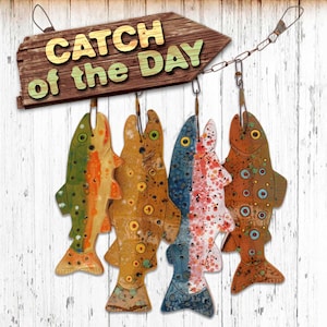 May include: Five wooden fish ornaments hanging from a sign that reads "Catch of the Day". The fish are painted in various colors and patterns, including green, orange, brown, blue, and red.