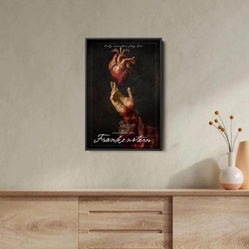 Op de afbeelding: Ingelijste kunstdruk met een menselijk hart boven een hand, met het woord "Frankenstein" in witte letters. Het kunstwerk heeft een donkere, gotische esthetiek, met de tekst "Only monsters play God" bovenaan.