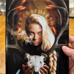 May include: A rectangular print featuring a blonde individual with blue eyes holding a white animal skull. Large dark antlers frame the figure, with glowing orange tips. A circular arrangement of runic symbols surrounds the head, and a signature is visible.