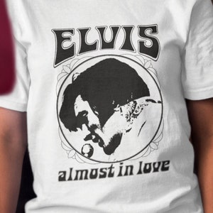 Puede incluir: Camiseta blanca con un gráfico en blanco y negro de Elvis Presley cantando. El gráfico incluye el texto "ELVIS" y "almost in love".