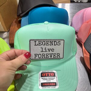May include: A stack of trucker hats in various colors, including black, teal, and mint green. The mint green hat features the text "LEGENDS live FOREVER" on a patch. The hats have a mesh back and a foam front.
