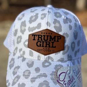 May include: White trucker hat with a grey leopard print pattern. The hat features a brown leather patch with the text "TRUMP GIRL GET OVER IT". The hat has a mesh back and a curved brim.